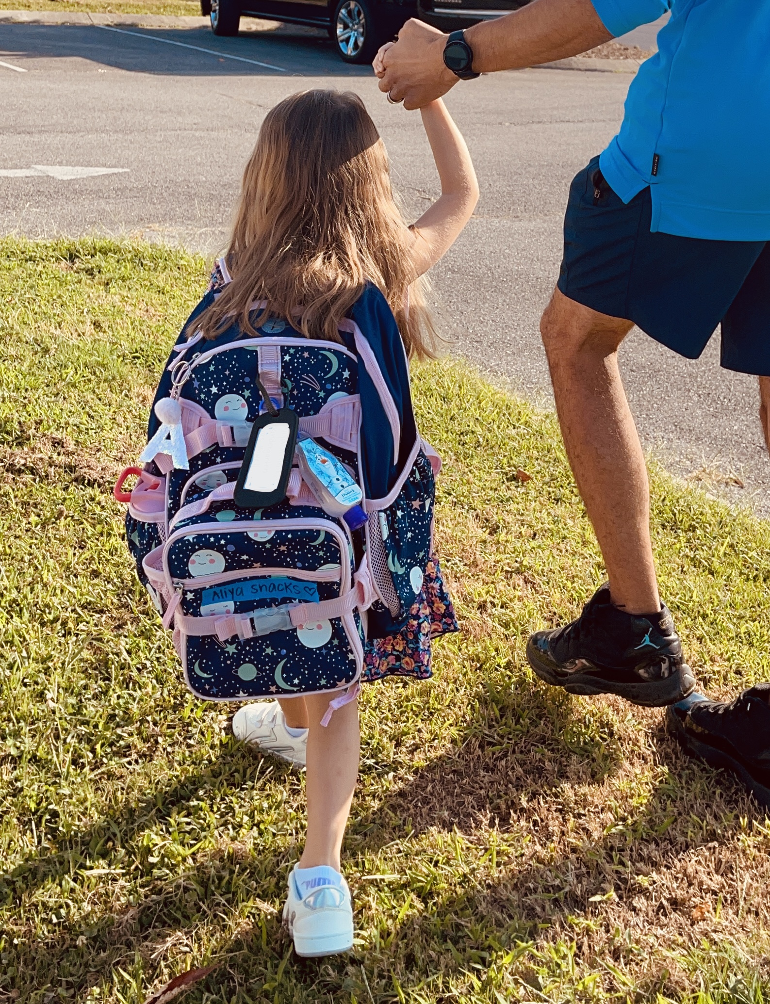 girl wearing a backpack and holding her dad's hand as she walks toward school on first day